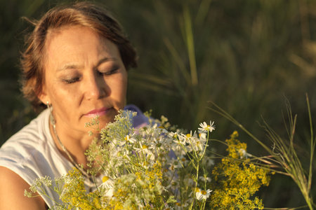 Enjoying a peaceful moment in nature, an older woman sits in a sunny meadow, cradling wildflowers in her hands, embodying the wisdom of age and the strength of self-acceptanceの写真素材