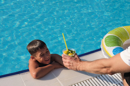Cheerful kid enjoying pool time, smiling brightly as an adult hand offers a tropical drink, emphasizing family, leisure, and sun-soaked momentsの写真素材