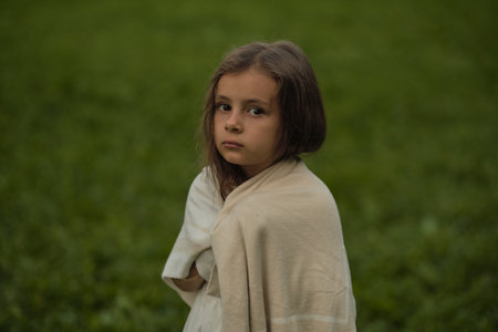 With an enigmatic gaze, a child stands cloaked in a blanket against a green field. This image speaks to the need for warmth and protection in a child s world.の写真素材