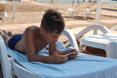 Kid on a chair engrossed in a smartphone, ocean ignored. Reflects the preference for digital interaction over natural exploration.の写真素材