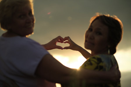 Two elderly women form a heart with their hands at sunset, symbolizing lasting friendship. The image resonates with themes of aging gracefully and maintaining bonds.の写真素材