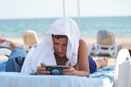 Boy with device on a sunbed, beach environment overlooked. Signals the growing concern of screen...