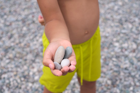 Young one offering rocks, vibrant shorts, soft beach setting. This image can symbolize the tactile learning experiences of youth.の写真素材