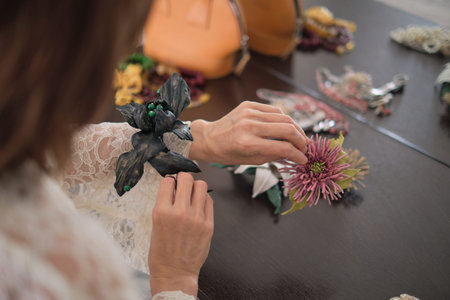 Hands delicately handle various floral accessories on a table, with an orange handbag in the...