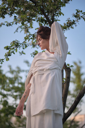 A woman dressed in white poses with her hands above her head, leaning against a tree. This scene reflects the calm and tranquility of being surrounded by nature.の写真素材