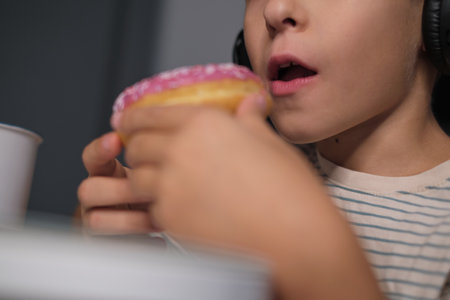 Close-up of a child holding and eating a donut with pink icing and sprinkles, showcasing the joy in the moment. Regular consumption of sugary foods can result in unhealthy weight gain and increase the risk of chronic diseases, underscoring the importance of balanced nutrition for children.の写真素材