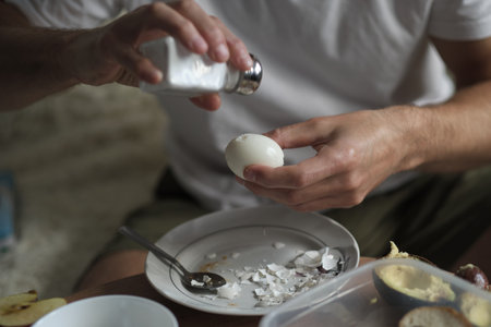A person adds salt to a freshly peeled hard-boiled egg, with broken eggshells scattered on a plate. This image captures a simple yet satisfying breakfast preparation.の写真素材