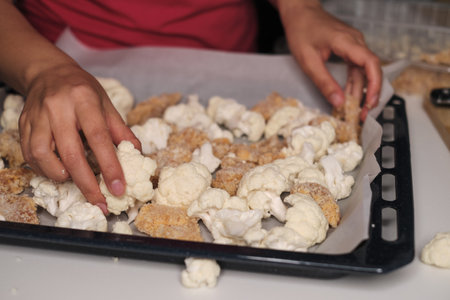Close-up of hands placing cauliflower and breaded chicken on a baking sheet, highlighting the meticulous preparation of a nutritious meal. This photo connects to the home-cooking movement and the focus on fresh produce.の写真素材