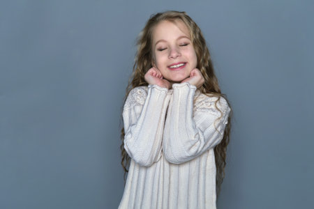 A young girl with long, wavy hair smiles sweetly, resting her hands gently near her face while wearing a cozy sweater. This image highlights the natural beauty of childrens hair and promotes the importance of gentle hair care.の写真素材