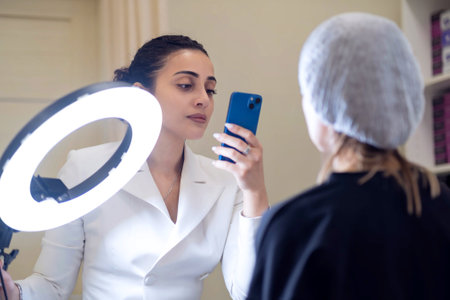 A beauty specialist documents her client s post-treatment progress with a smartphone and ring light. Capturing authentic results enhances trust in the ever-growing digital beauty market.の写真素材