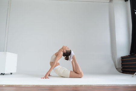 Dressed in a beige sports outfit, a woman bends backward in a graceful yoga pose against a plain...