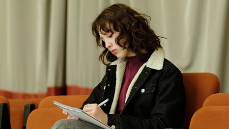 A girl pens her thoughts in a notebook while seated in a serene indoor space. The photo emphasizes...
