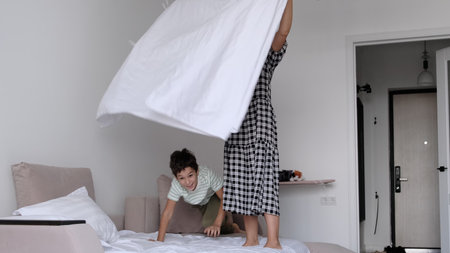 A woman tosses a blanket into the air as her son jumps onto the bed, both immersed in playful...