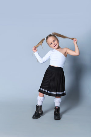 joyful young girl pulls on her pigtails while smiling, dressed in a cute and playful outfit. The black pleated skirt, white top, and lace socks reflect a mix of fashion and fun, capturing the essence of a carefree childhood moment.の写真素材