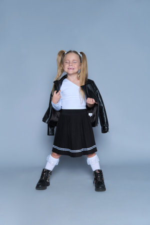 A confident portrait of a young girl, dressed in a modern outfit with a leather jacket, black boots, and pleated skirt. Her bold stance and playful expression make this a perfect shot for childrens fashion, lifestyle, or portrait photography.の写真素材