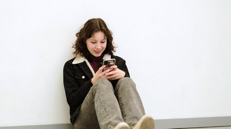 A seated girl with a happy, thoughtful expression holds her phone, enjoying a quiet moment. This image reflects themes of connection, technology, and the small joys of modern life.の写真素材
