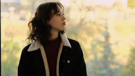 A young girl stands by a window, gazing thoughtfully at the bright outdoor scenery. This image captures themes of introspection, connection to nature, and quiet reflection.の写真素材