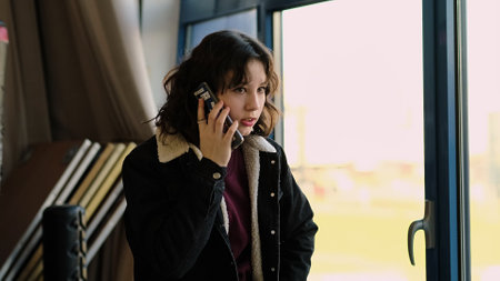 A girl casually holds a smartphone to her ear near a sunlit window, her gaze directed outward. The photo emphasizes personal communication and the simplicity of daily moments.の写真素材
