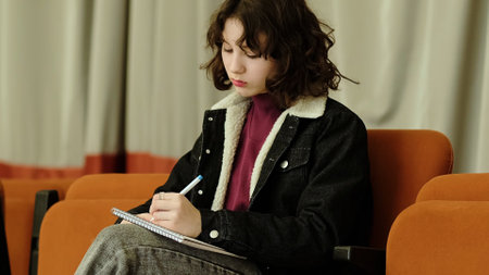A focused girl sits in an auditorium chair, immersed in her notebook writing. The scene captures the dedication and introspection involved in personal growth and creativity.の写真素材