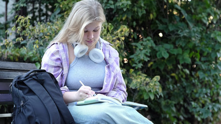 A teenage girl studies outdoors, pen in hand and books open, with a backpack beside her. This photo captures a peaceful moment of learning in a natural setting.の写真素材