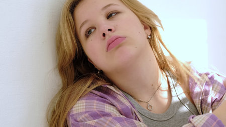 A young woman leans against a wall, gazing thoughtfully with a subtle expression of sadness. The...