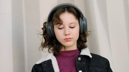 A young girl with headphones looks down thoughtfully, immersed in a quiet moment. This image captures introspection, focus, and the personal connection with audio or music.の写真素材