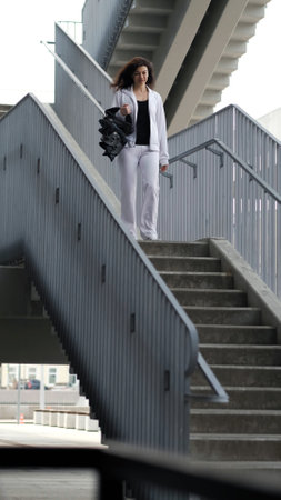 A woman dressed in sportswear moves down a metal staircase, carrying specialized jumping boots. The industrial backdrop emphasizes the fusion of fitness, architecture, and city movement.の写真素材