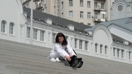 A woman in a white tracksuit sits on urban stairs, fastening her Kangoo Jumps rebound shoes. The fusion of modern fitness and classical architecture symbolizes the blend of active living and city aesthetics.の写真素材