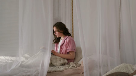 A young woman sits in a serene bedroom surrounded by white curtains, deeply focused on a book. This image highlights the importance of finding tranquility in a busy, modern world.の写真素材