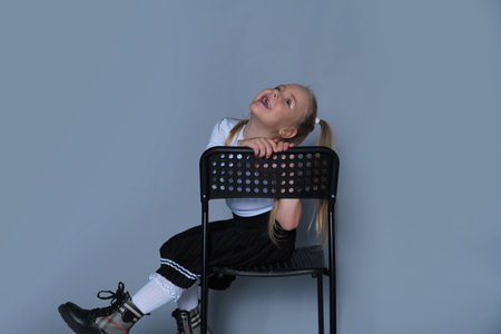 A playful 5-year-old girl is photographed in a studio, sitting on a black chair with a big, joyful expression. Dressed in a stylish one-shoulder top and black pleated skirt, she radiates energy and fun, perfect for her modeling portfolio.の写真素材