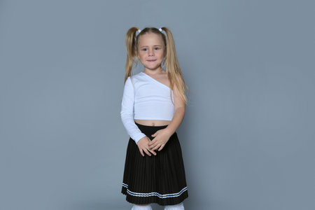 A young girl with blonde pigtails and a gap-toothed smile wears a white one-shoulder top and a black pleated skirt. The photo showcases her playful and carefree spirit, capturing a moment of childhood joy.の写真素材