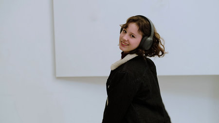 A happy young girl enjoys listening to music, moving gracefully in a bright, simple space. This...