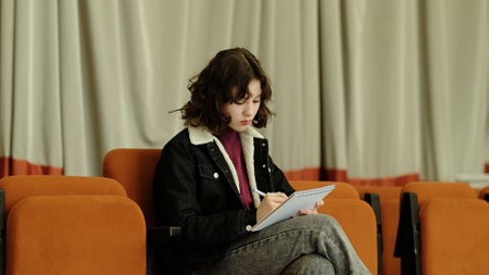 A young girl focuses on her writing while seated in an auditorium chair, engrossed in her notebook. This image reflects themes of creativity, learning, and thoughtful concentration.の写真素材