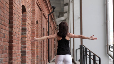 A woman in sportswear stretches her arms wide while standing near a brick wall. The combination of movement and urban architecture reflects the harmony between strength, flexibility, and modern city life.の写真素材