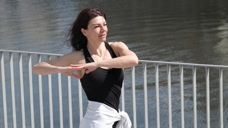 A woman in sportswear moves gracefully near a river, her arms extended in a dynamic pose. The connection between movement and nature reflects the modern trend of outdoor fitness and mindful exercise.の写真素材