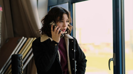 A young girl holds a smartphone to her ear, gazing thoughtfully out of a window. This image captures themes of modern communication, contemplation, and youthful connection.の写真素材