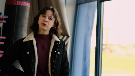 A confident young girl stands indoors with natural light streaming in, her expression calm and poised. The photo symbolizes individuality, self-expression, and youthful resilience.の写真素材