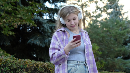 A young woman with headphones and a plaid shirt looks at her phone while enjoying an outdoor moment. The photo highlights the blend of technology and relaxation in nature.の写真素材
