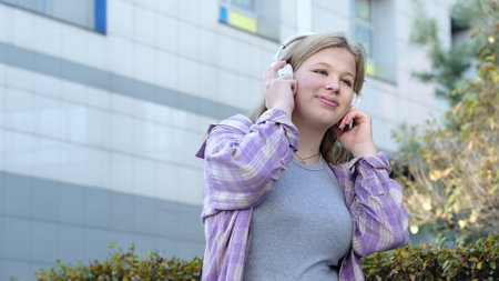 A blonde girl in a plaid shirt enjoys her music while standing near a contemporary building. The image highlights the harmony between individuality and modern surroundings.の写真素材