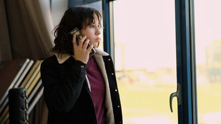 A girl speaks on her phone while standing near a bright window, her expression calm and focused. The photo reflects themes of technology, connection, and real-time interaction.の写真素材