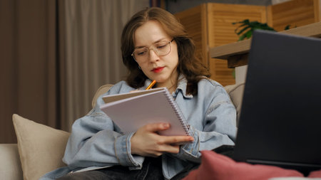Engrossed in her work, a woman writes in a notebook while using a laptop. This image reflects the flexibility and comfort of remote work setups in modern homes.の写真素材