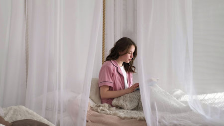 A thoughtful woman reads a book under delicate curtains, creating a serene atmosphere. It highlights the modern need for introspection and finding peace in everyday life.の写真素材