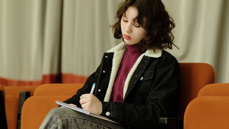 A young girl with a pen in hand attentively works in her notebook while seated in orange theater seating. This image portrays themes of education, mindfulness, and planning.の写真素材