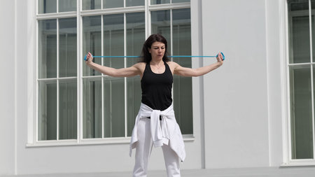A woman performs a resistance band exercise, engaging her upper body muscles outdoors. The image...