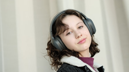 A thoughtful girl with headphones relaxes in a light-filled space, her expression serene. The image...