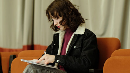 A girl writes attentively in her notebook while seated indoors, framed by a neutral curtain...
