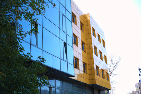 Modern office building with bright yellow facade panels and mirrored glass windows reflecting a nearby high-rise tower. The image illustrates the use of ventilated facades and reflective glazing in contemporary architecture, suitable for topics about modern construction materials, energy-efficient design, or urban redevelopment.の写真素材