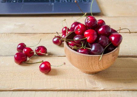 Red berries in a wooden bowl. The cherries are on a wooden table, behind a laptop. Fruits lie on a wooden background, daylight.の写真素材