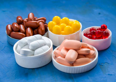 A group of vitamins to support health. Multi-colored dietary supplements lie on different sizes of lids, on a blue background.の写真素材