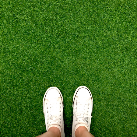Feet in white sneakers on green grass, top view, informal style great for any use.の写真素材